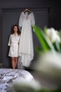 bride in a silk robe next to a white wedding dress hanging on the closet.