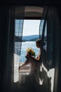 The bride sits at an open window with a sea view and fluttering curtains and holds a wedding bouquet with orange flowers Royalty Free Stock Photo