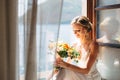 The bride sits at an open window with a sea view and fluttering curtains and holds a wedding bouquet with orange flowers Royalty Free Stock Photo