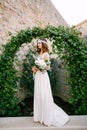 A bride stands at a graceful arch entwined with wild grapes in the old town of Budva and holds a bouquet in her hands Royalty Free Stock Photo