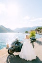 A bride in sunglasses and a wedding dress stands near a motorcycle on the pier in the old town of Perast