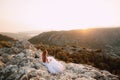 The bride in tender wedding dress sits on the rock on Mount Lovcen and looks at the Bay of Kotor