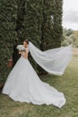 the bride in a wedding dress with a long train and a veil holds a wedding bouquet of roses against the background of tall green Royalty Free Stock Photo