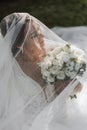 a bride in a wedding dress with a long train and a veil holds a wedding bouquet of roses while sitting under a veil Royalty Free Stock Photo