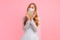Bride in a wedding dress with a medical protective mask on her face, holding a wooden heart on a pink background. Quarantine, Royalty Free Stock Photo