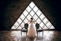 The bride in a white wedding dress stands near two striped armchairs, against the background of a triangular design window Royalty Free Stock Photo