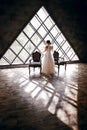The bride in a white wedding dress stands near two striped armchairs, against the background of a triangular design window Royalty Free Stock Photo