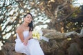 Bride in the wedding dress sits on the roots of a tree in the middle of the stones on the background of tropical trees