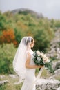 Bride in a white dress with a veil holds a bouquet of flowers in her hands against a background of greenery and Royalty Free Stock Photo