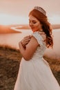 Bride in white wedding dress with a crown on her head stands on cliff against the background of the river and islands