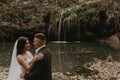 bride in white wedding dress and long veil hugs groom in a suit small pond lake river waterfall Burbun Royalty Free Stock Photo