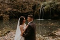 bride in white wedding dress and long veil hugs groom in a suit small pond lake river waterfall Burbun Royalty Free Stock Photo