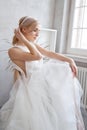 Bride woman in a light summer wedding dress sitting at the window and waiting for the wedding ceremony. Girl with perfect hair and