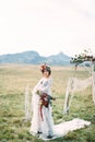 Bride in a wreath with a bouquet stands on a white carpet in front of a wedding arch in a meadow