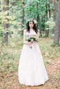 The bride with wreath on head in a stylish wedding dress in nature forest with a rustic bouquet. Photo shoot in pine