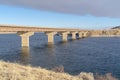 Bridge across blue lake with views of snow covered hills and cloudy blue sky