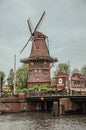 Bridge over canal, trees and old windmill on the bank with cloudy sky in Amsterdam. Royalty Free Stock Photo
