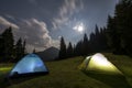 Bright big moon in dark blue cloudy sky over two tourist tents on green grassy forest clearing among tall pine trees on distant Royalty Free Stock Photo