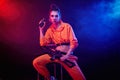 A bright brunette in an orange jumpsuit with a machine gun on a dark background in neon light. talking on the radio Royalty Free Stock Photo