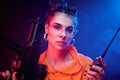 A bright brunette in an orange jumpsuit with a machine gun on a dark background in neon light. talking on the radio Royalty Free Stock Photo