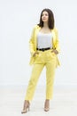 Confident brunette girl holding hands in pockets of bright stylish yellow suit and posing in studio on white background. Royalty Free Stock Photo
