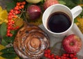 A A bright composition with a cup of strong black tea, a sweet bun with raisins, ash berries, apples and colorful autumn leaves on Royalty Free Stock Photo