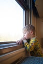 A bright, cute three-year-old boy riding a train looks out the window, behind which a dull landscape sweeps through