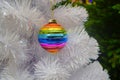 Bright glass rainbow colored Christmas ball, bauble hanging on a white artificial christmas tree. On background of green christmas