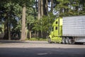Bright green big rig industrial semi truck with refrigerator semi trailer running on the rest area parking lot with old trees to Royalty Free Stock Photo