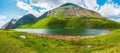 Bright green blue alpine landscape with mountain lake in highland valley in sunlight and big mountain under blue cloudy sky. Royalty Free Stock Photo