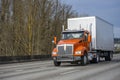 Bright orange day cab big rig semi truck with box trailer running on the wide multiline highway road with trees on the side