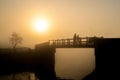 Bright orange misty sunrise as couple silhouette walk over bridge looking at view in fog. Wooden footbridge crossing river canal