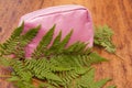 Bright pink cosmetic bag with spakles covered with green fern leaves on a wooden background