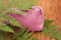 Bright pink cosmetic bag with spakles covered with green fern leaves on a wooden background