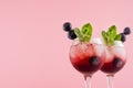 Bright red alcoholic shots in two wineglasses with blueberry, ice cubes, green mint closeup, top section on pastel pink color.