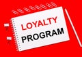 On a bright red background, a white pencil, white paper clips, and a white notebook with the text LOYALTY PROGRAM