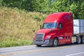 Bright red bonnet big rig semi truck transporting commercial cargo on flat bed semi trailer running on the road with hillside Royalty Free Stock Photo
