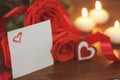 Three bright red roses, sign heart, satin ribbon, sheet of paper and burning candles on wooden background closeup.