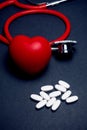 Bright red stethoscope and heart and pile of white pills on a dark blue surface, Health and treatment of heart related disease