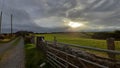 Bright sun in cloudy sunset sky shining over lush green fenced field in the countryside