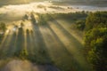 Bright sun rays through trees on misty meadow near river. Scenic spring landscape. Beautiful vivid sunbeams in morning