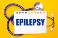 On a bright yellow background, a blue stethoscope and a sheet of paper with the text EPILEPSY. Medical concept