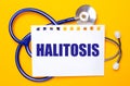 On a bright yellow background, a blue stethoscope and a sheet of paper with the text HALITOSIS. Medical concept