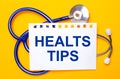 On a bright yellow background, a blue stethoscope and a sheet of paper with the text HEALTS TIPS. Medical concept