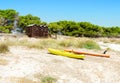 A bright yellow kayak and an orange board lie on a sandy beach next to wooden cabanas against a backdrop of green pine trees and a Royalty Free Stock Photo