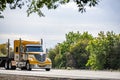 Stylish big rig yellow semi truck with chrome accessories transporting step down semi trailer moving on the road with trees on the Royalty Free Stock Photo