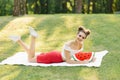 A bright and young girl lies on a white plaid on a lawn in a park with a fan hairstyle and bright makeup, holds a watermelon pillo