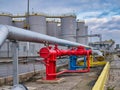 Brightly coloured industrial pipelines, control valves and a low yellow wall with a background of large, tall grey storage tanks