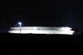 Brightly lit large white industrial oil refinery tank with protective fence placed on top of small hill on warm summer night