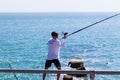 Brighton, UK - June 2018 Man with Cap Standing on Shore Holding Sideways Long Fishing Rod with Reel. Crystal Blue Wavy